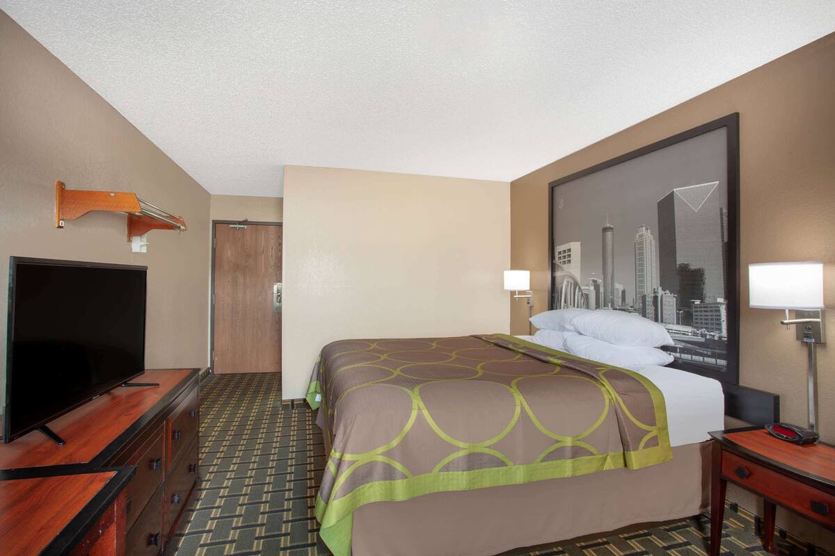 A hotel room with a bed, a television on a wooden dresser, a cityscape poster on the wall, and a door.