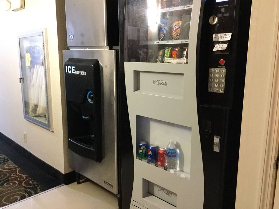 A vending machine with a push button and a coin dispenser, located in a hallway with a patterned carpet.