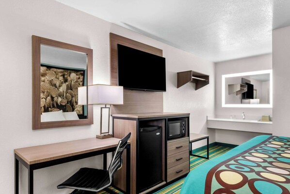Habitación de hotel con cama, escritorio con silla, televisor, microondas, refrigerador y baño con espejo.