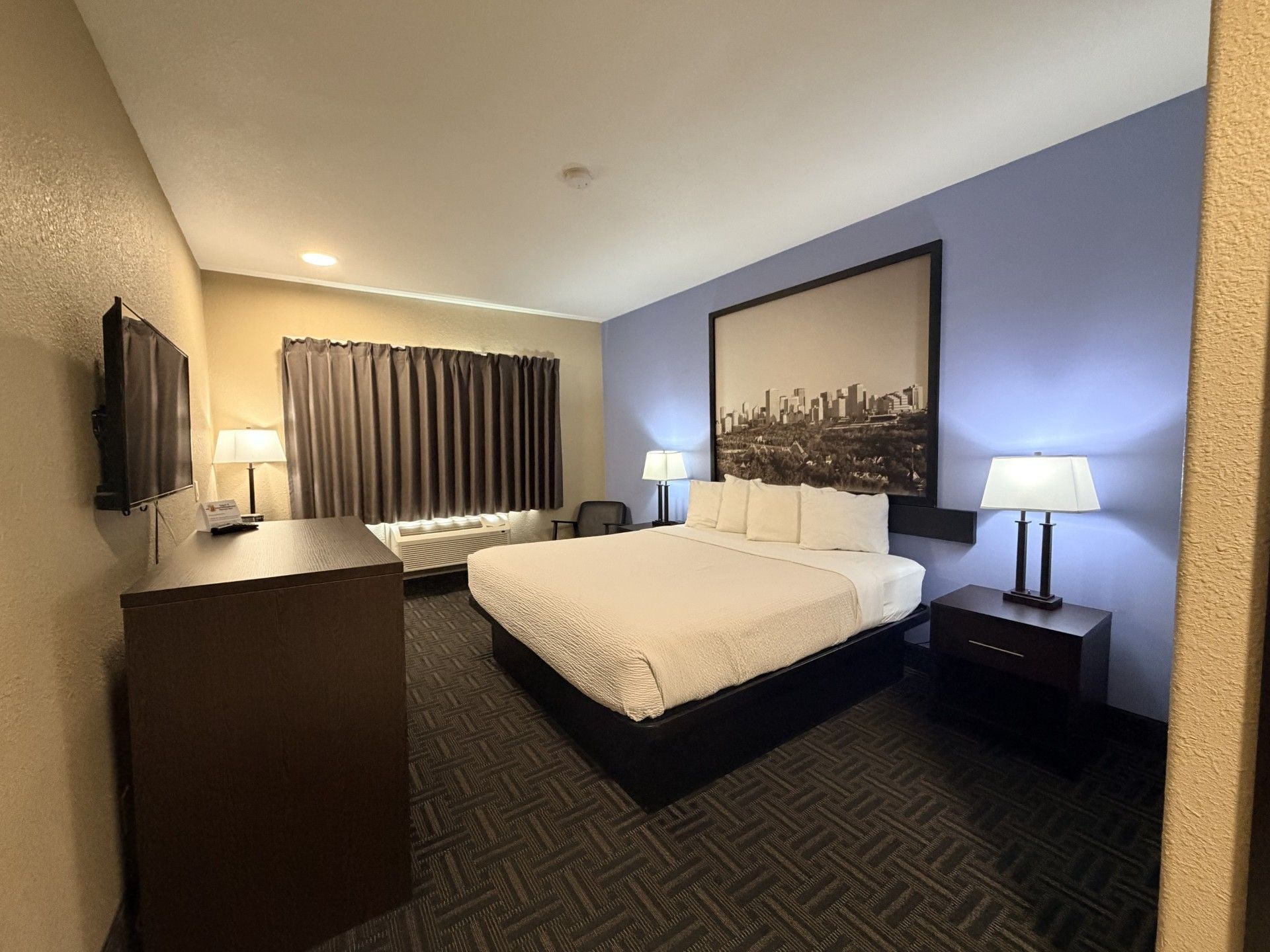 A hotel room with a bed, two bedside tables, a desk, a chair, a television, and a cityscape painting on the wall.