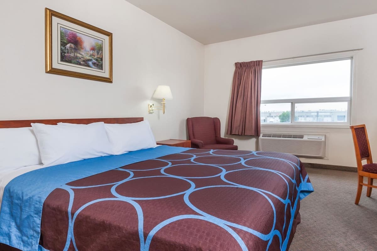 A hotel room with a bed, a chair, a desk, a window with curtains, a wall-mounted lamp, and a framed picture on the wall.