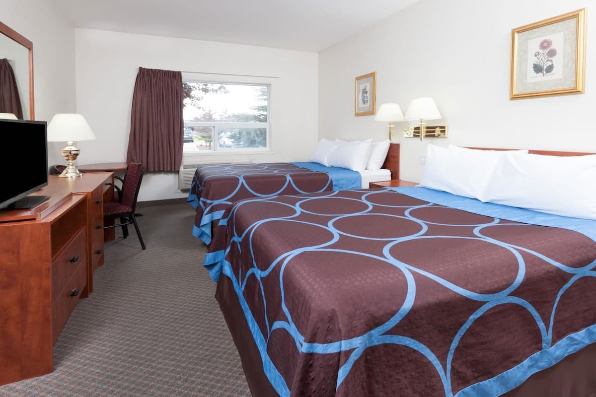 A hotel room with two beds, a desk with a TV, a chair, a window with curtains, and wall-mounted lamps.