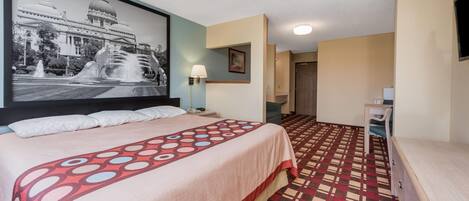 A hotel room with a bed, a large wall-mounted picture, a patterned carpet, a desk, and a chair.
