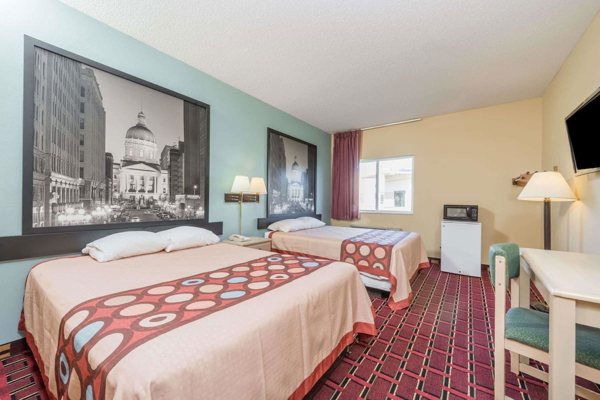 Habitación de hotel con dos camas, un escritorio, un televisor y un cuadro grande con un paisaje urbano.