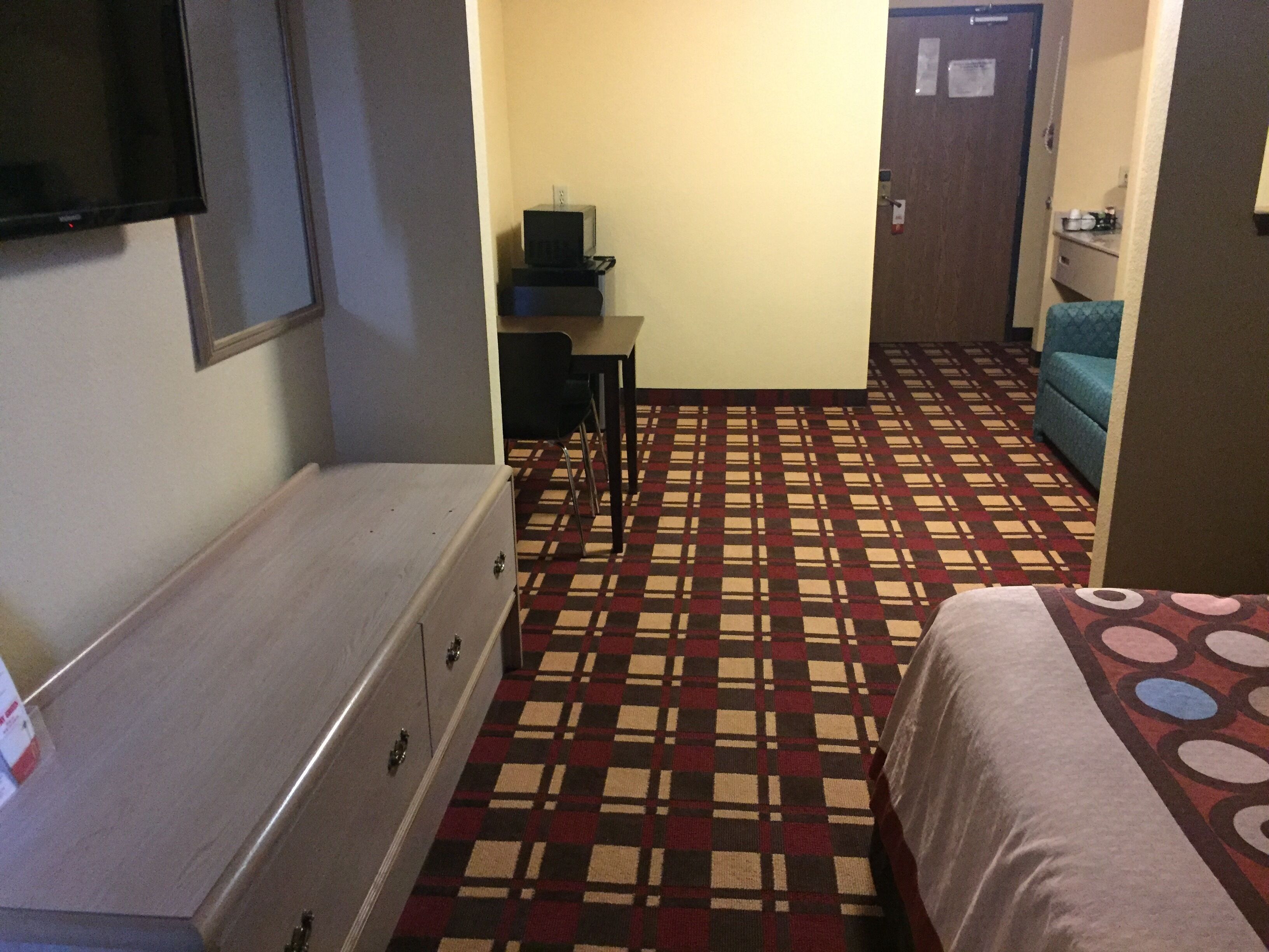 A hotel room with a bed, a dresser, a desk with a chair, a television, and a patterned carpet.