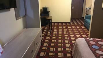 A hotel room with a bed, a dresser, a desk with a chair, a television, and a patterned carpet.