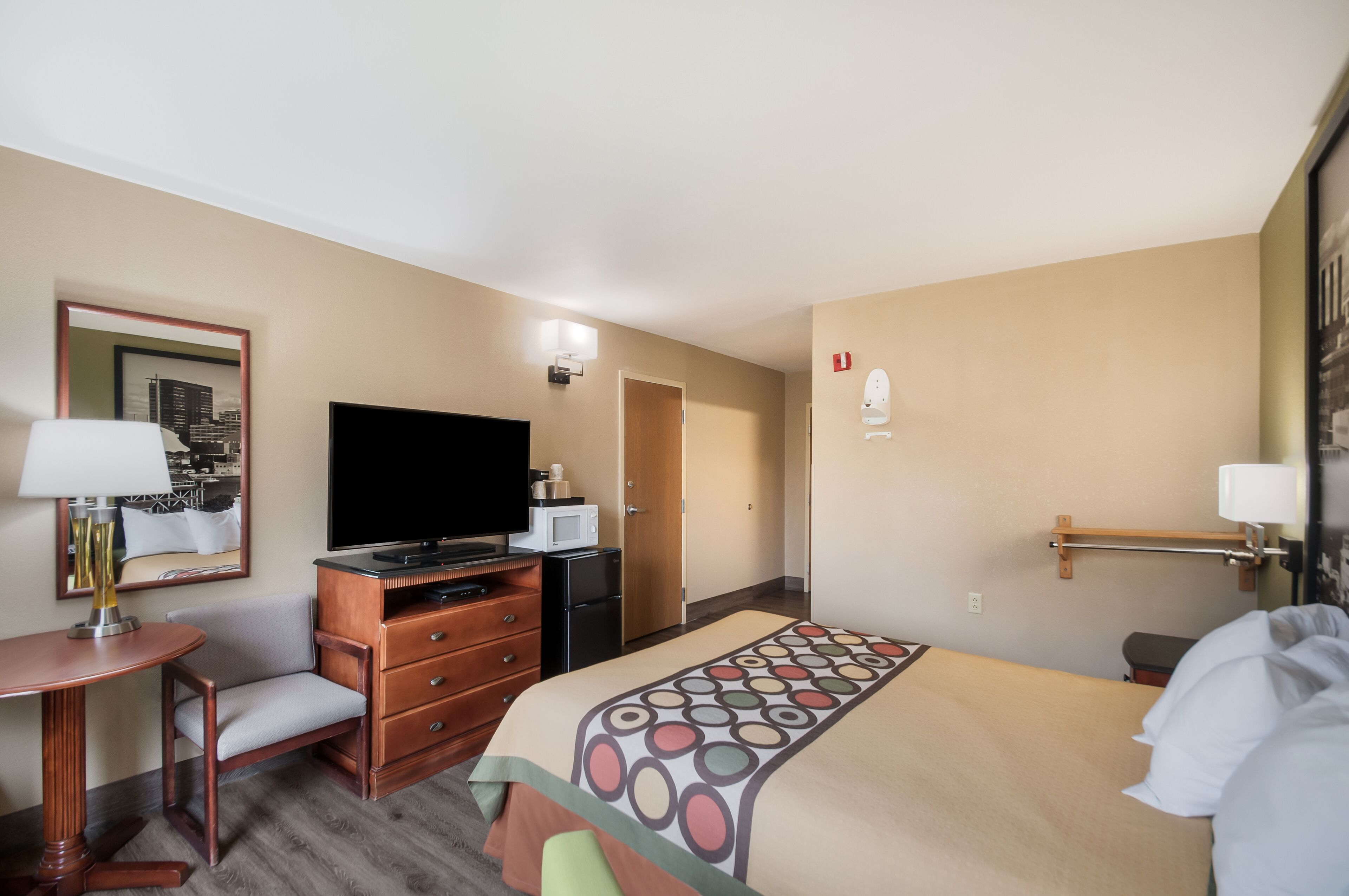 Habitación de hotel con cama, televisor, escritorio con lámpara, cómoda y puerta que da a otra habitación.