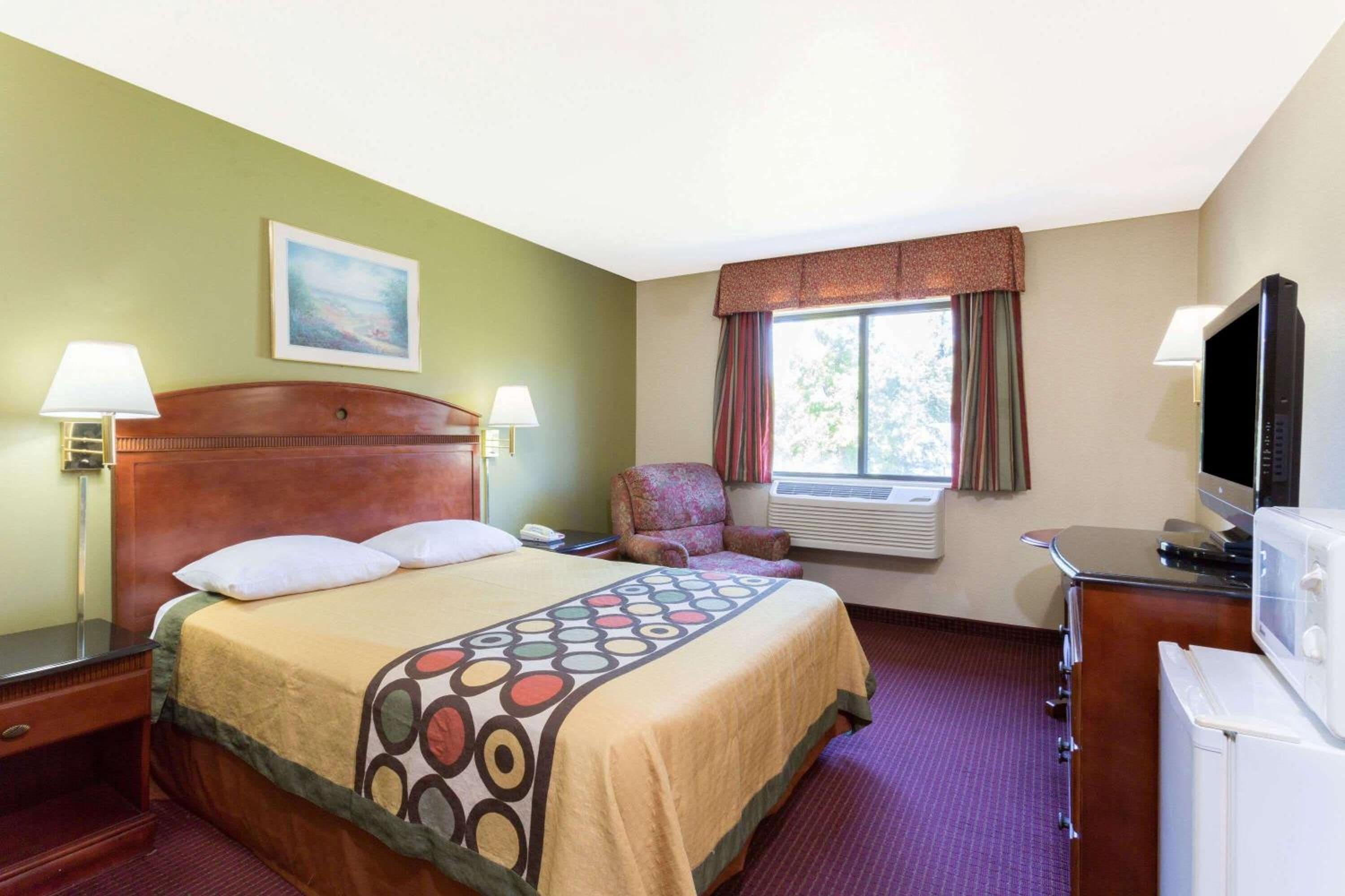 Ein Hotelzimmer mit Bett, Sessel, Schreibtisch mit Fernseher und Fenster mit Vorhängen.