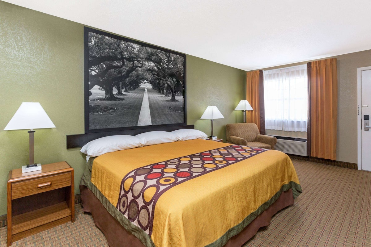 Een hotelkamer met een groot bed, twee bedlampen, een stoel, een deur en een groot ingelijst schilderij aan de muur.