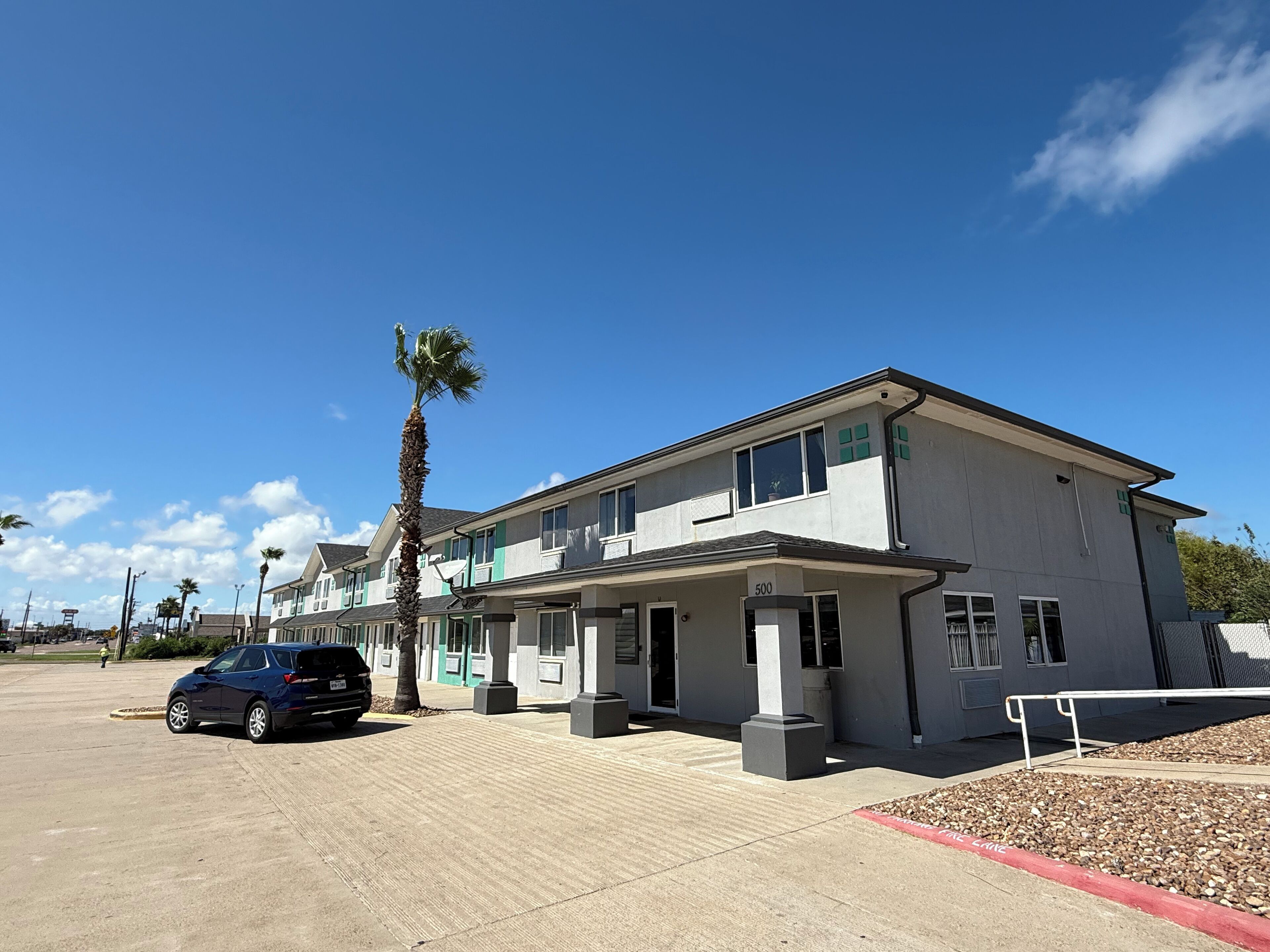 A multi-story building with a parking lot, a palm tree, and a blue car.