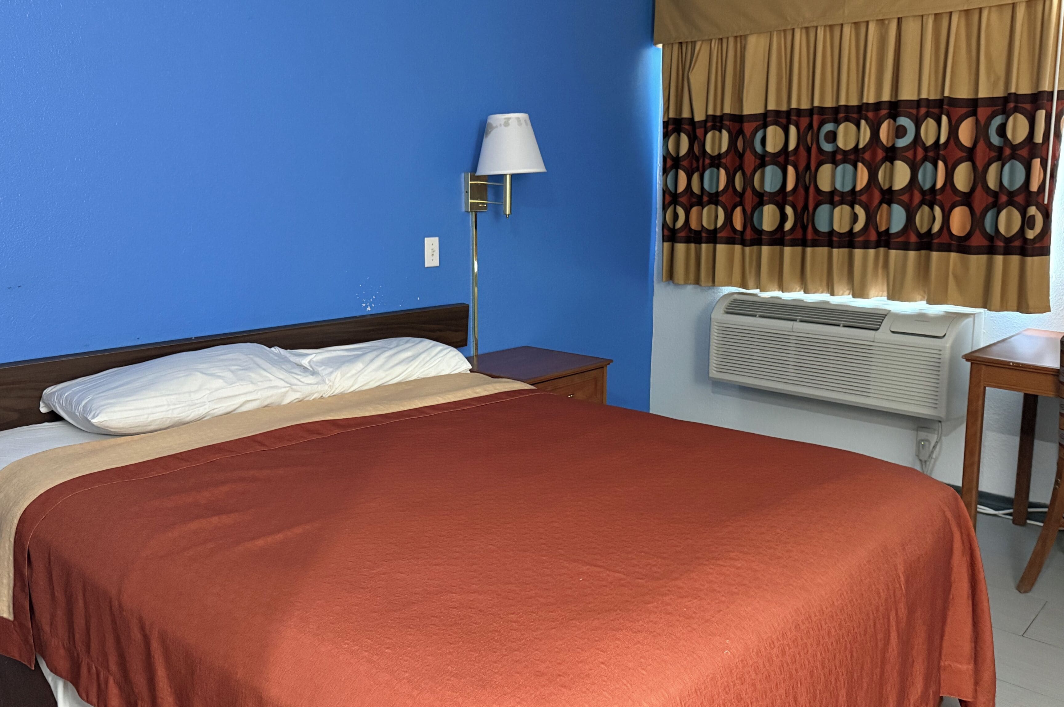 Habitación de hotel con cama, mesita de noche, lámpara, equipo de aire acondicionado y ventana con cortinas.