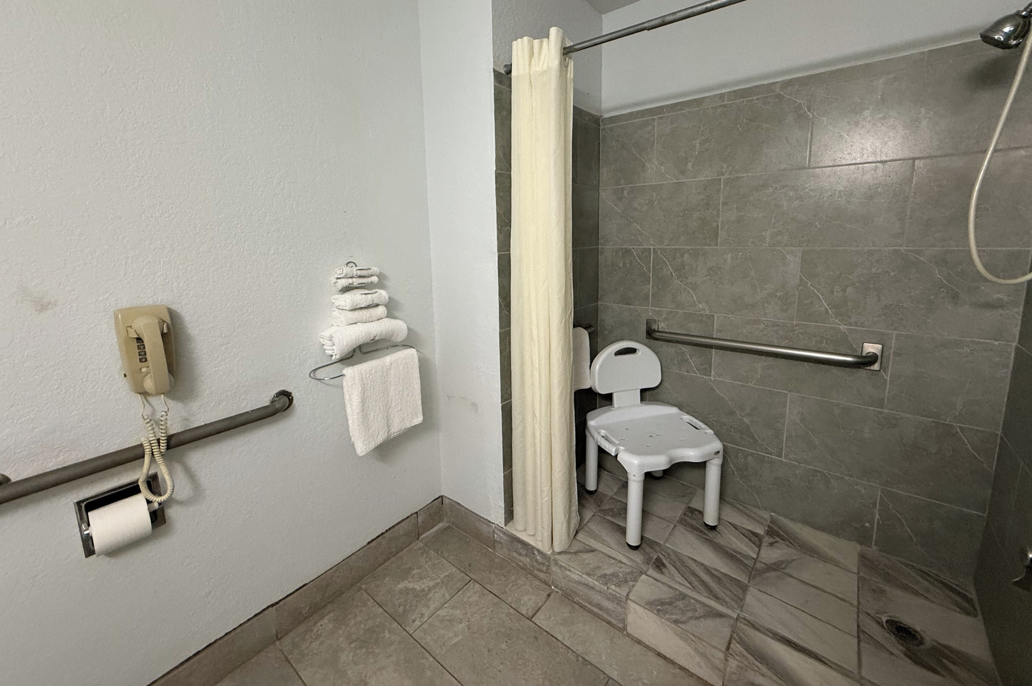 Une salle de bain équipée d’une douche, de toilettes, d’une barre d’appui, d’un téléphone et d’une petite chaise blanche.