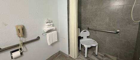 Une salle de bain équipée d’une douche, de toilettes, d’une barre d’appui, d’un téléphone et d’une petite chaise blanche.