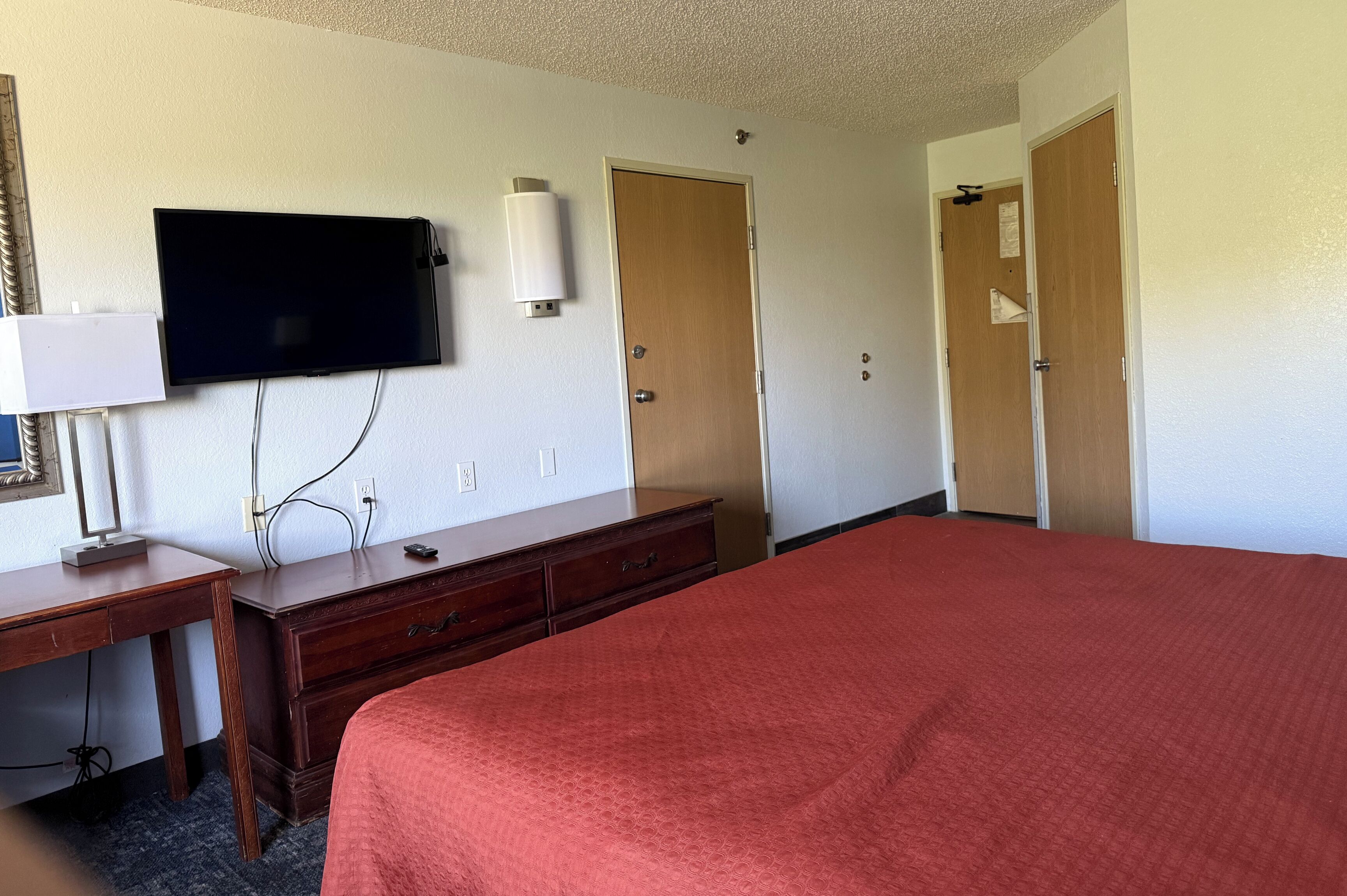 A hotel room with a bed, a desk, a television, and two doors.