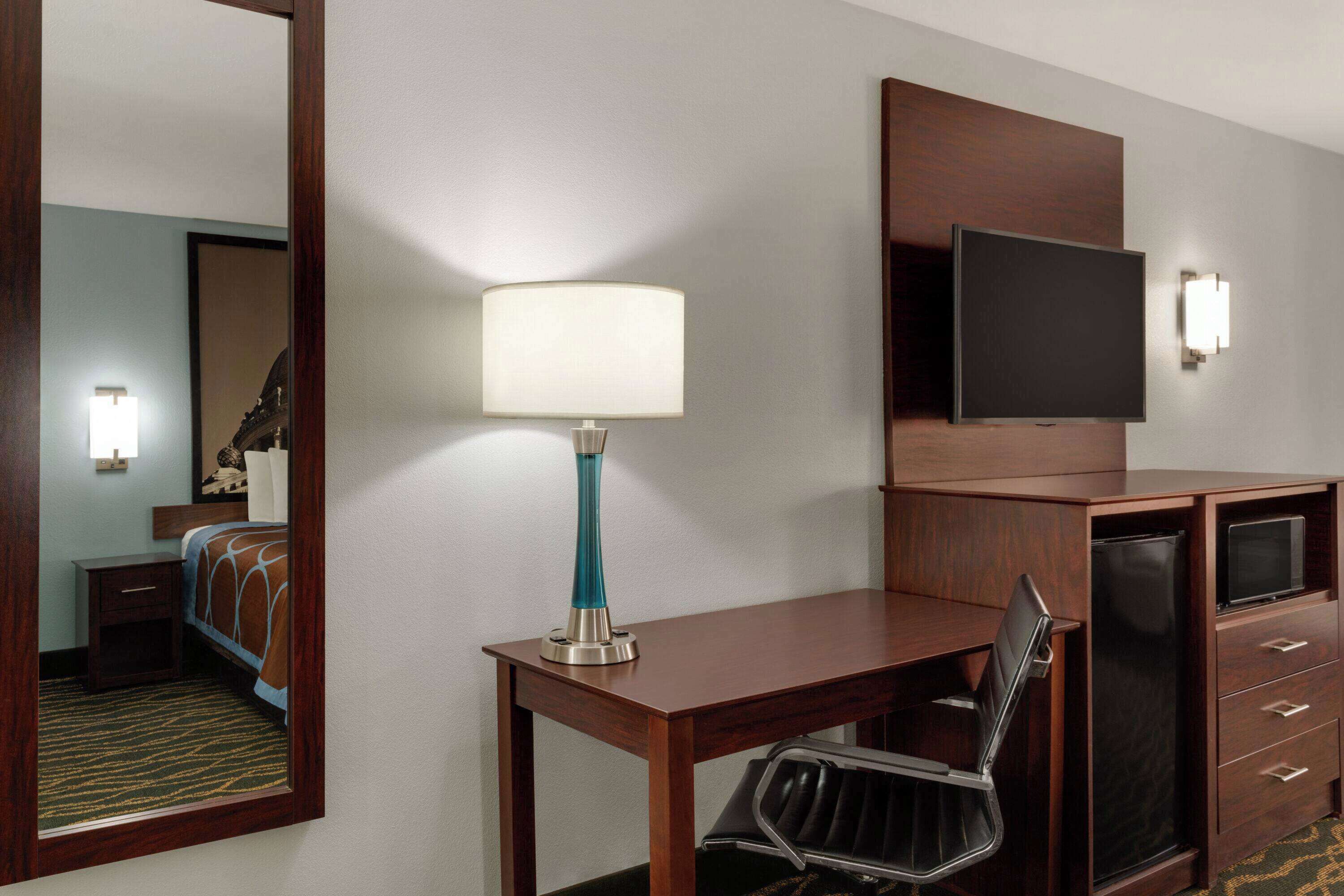 Quarto de hotel com escrivaninha de madeira, cadeira, televisão fixada na parede, abajur e cama com cabeceira.