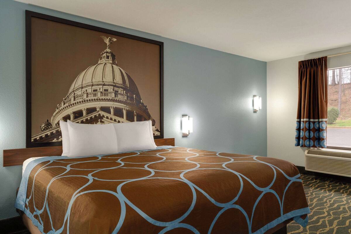 Een hotelkamer met een groot bed, een wandlamp, een raam met gordijnen en een inlijsting foto van een gebouw.