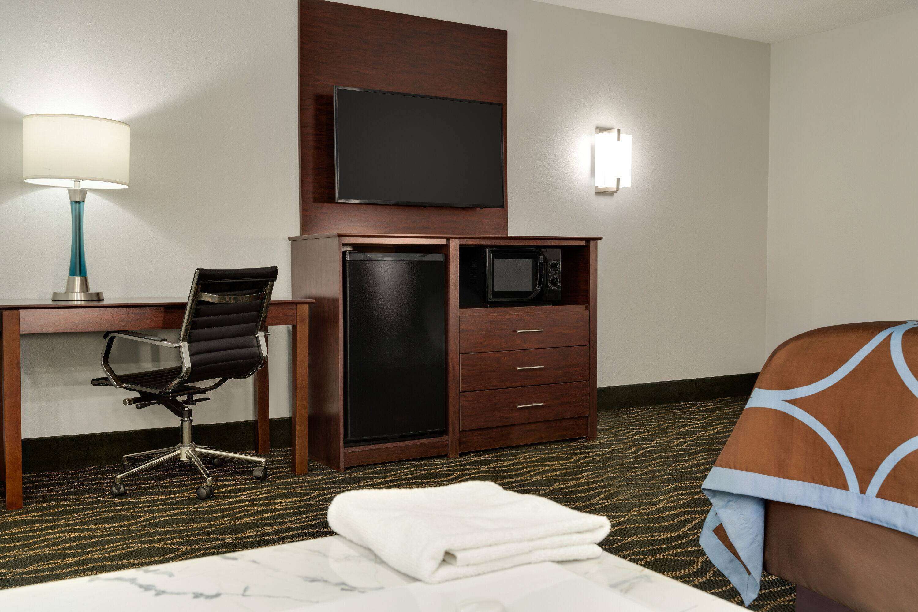 A hotel room with a desk, chair, TV, microwave, and a bed with a blanket.
