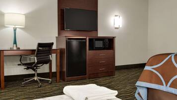 A hotel room with a desk, chair, TV, microwave, and a bed with a blanket.