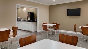A modern hotel room with a kitchenette, a flat-screen TV mounted on the wall, and a dining area with white tables and brown chairs. - Super 8 by Wyndham Pearl/Jackson Airport (Pearl)