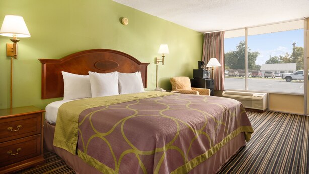 A hotel room with a large bed, two bedside lamps, a chair, and a window with a view of the parking lot. - American INN of Orangeburg (Orangeburg)