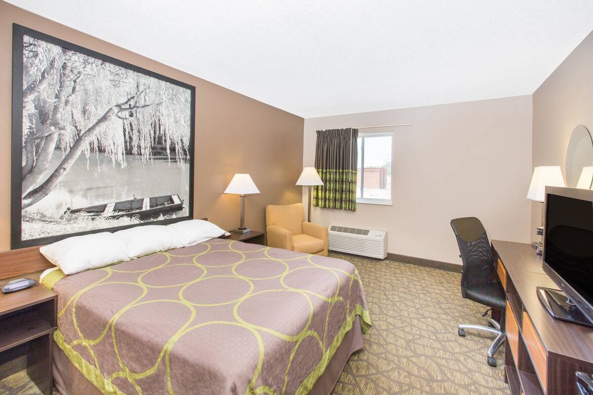 A hotel room with a bed, a desk with a computer, a chair, a TV, and a large window with curtains.