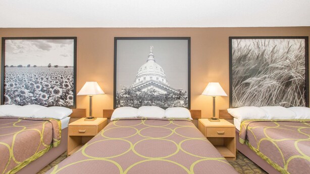 Quarto de hotel com duas camas, cada uma com colcha estampada e uma luminária de cada lado. Há três quadros na parede, um de um campo, um de um edifício e um de grama alta.