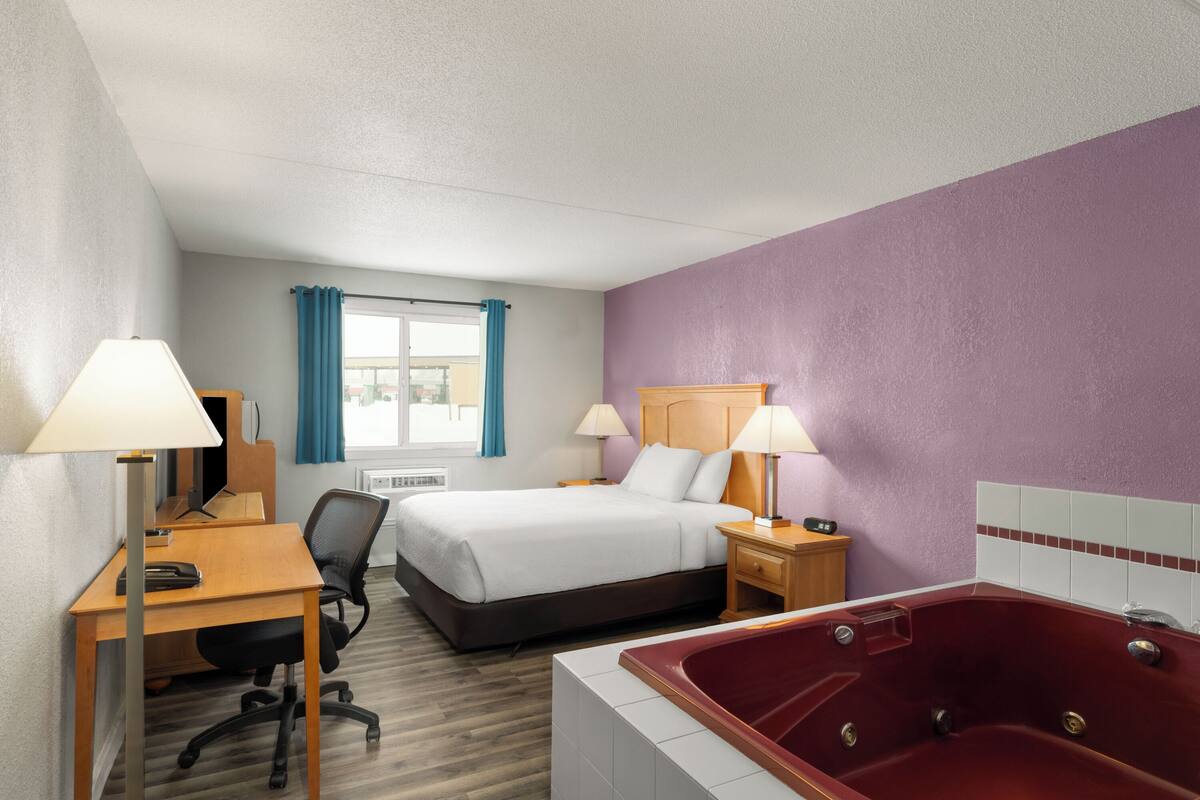 Habitación de hotel con cama, escritorio, silla, dos lámparas, ventana con cortinas y una bañera de hidromasaje roja.