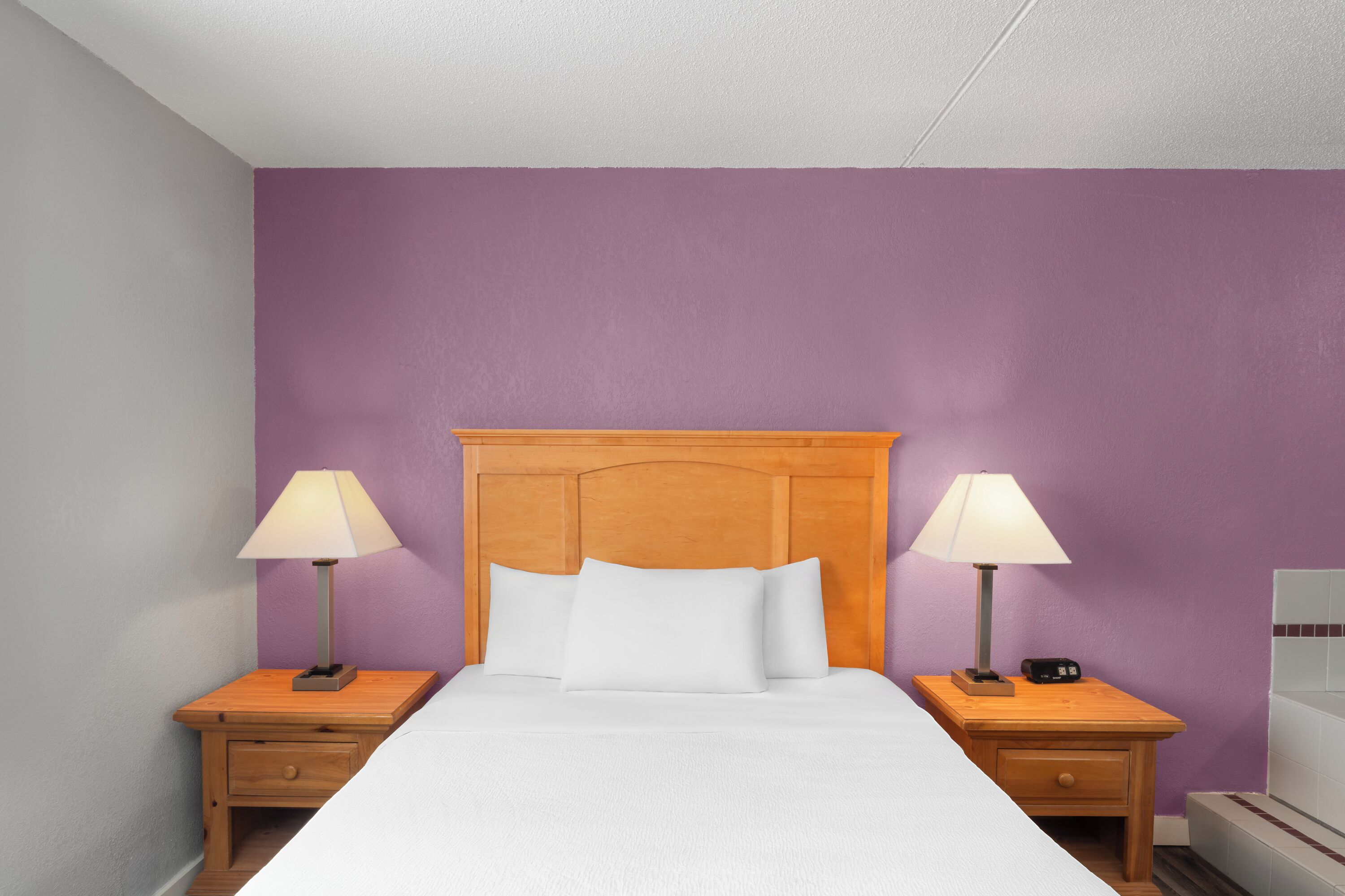 A neatly made bed with white bedding, flanked by two wooden nightstands with lamps, in a room with purple walls.