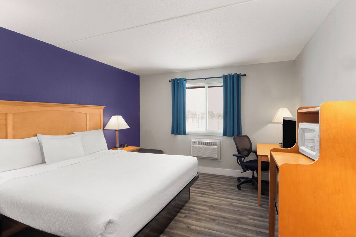 Habitación de hotel con una cama grande, un escritorio con ordenador, una silla, una lámpara y un ventanal con cortinas.