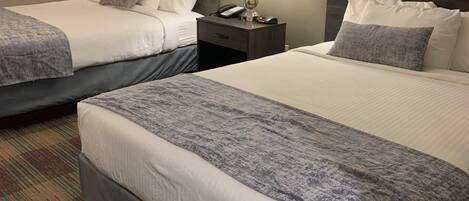 A hotel room with two beds, each with a grey and white duvet, a small bedside table with a lamp, and a patterned carpet.