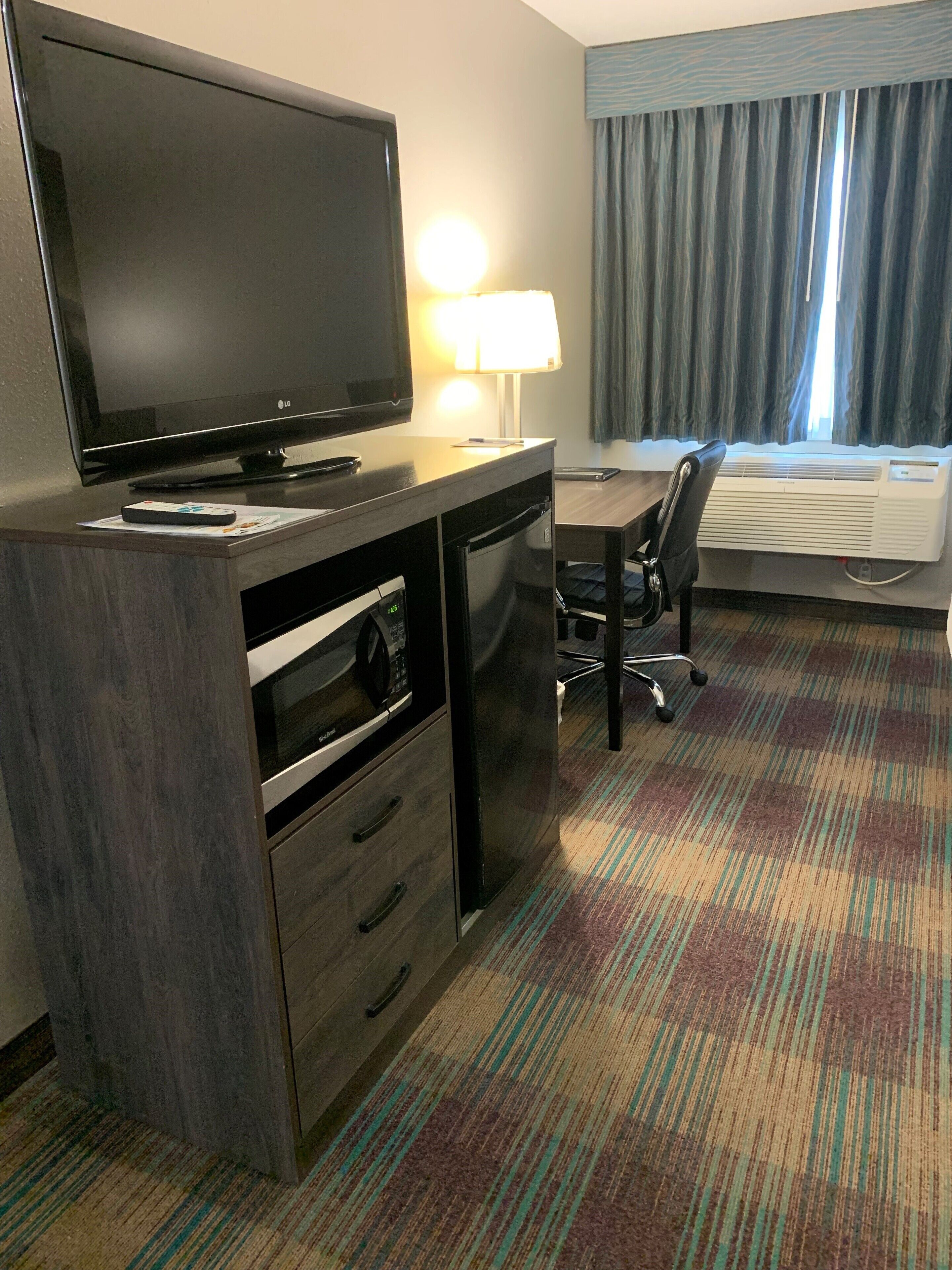 A hotel room with a flat-screen TV, a desk with a chair, a lamp, an air conditioner, and a patterned carpet.