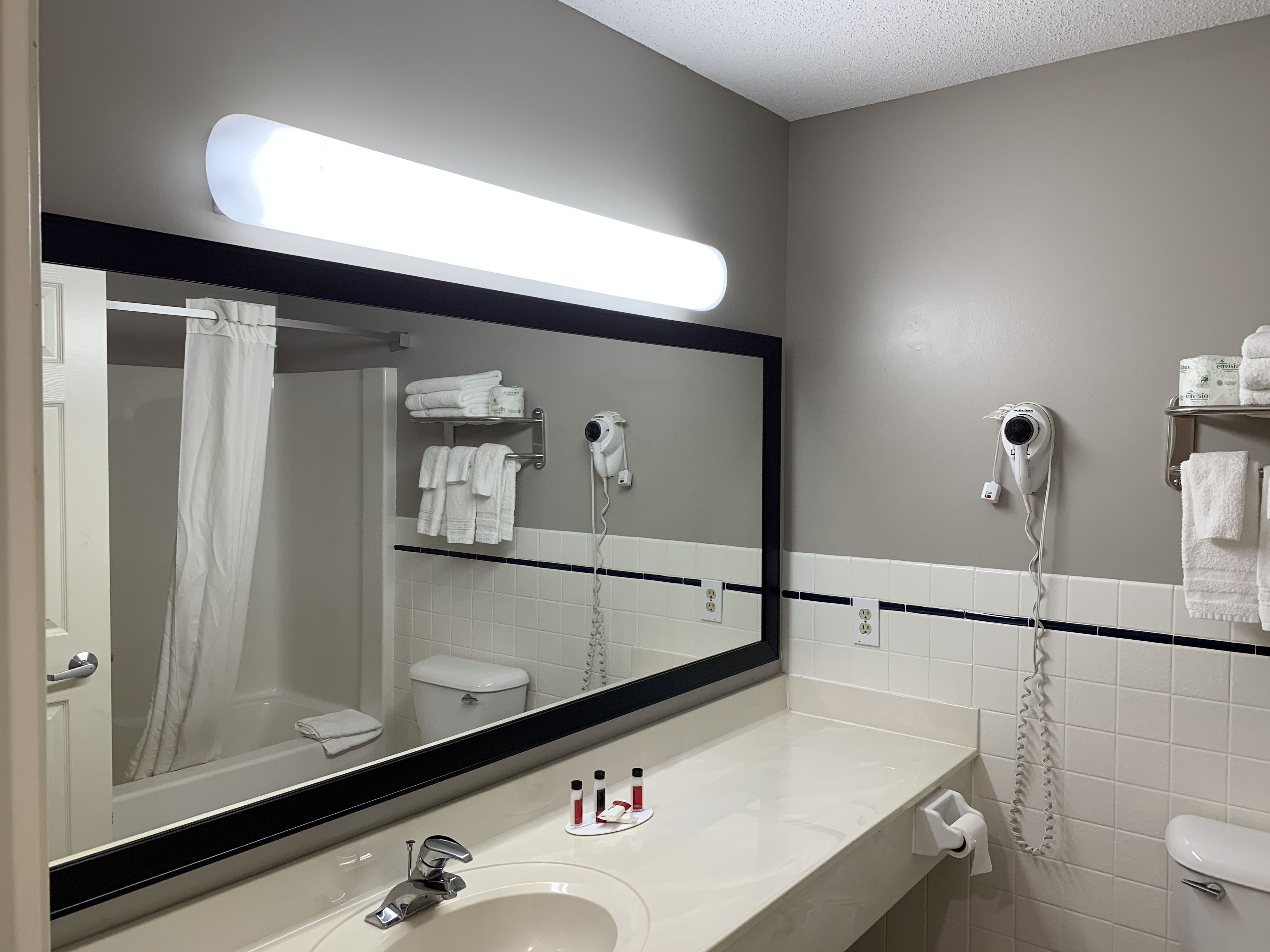 Une salle de bain avec un grand miroir, un lavabo, un porte-serviettes et deux sèche-cheveux.