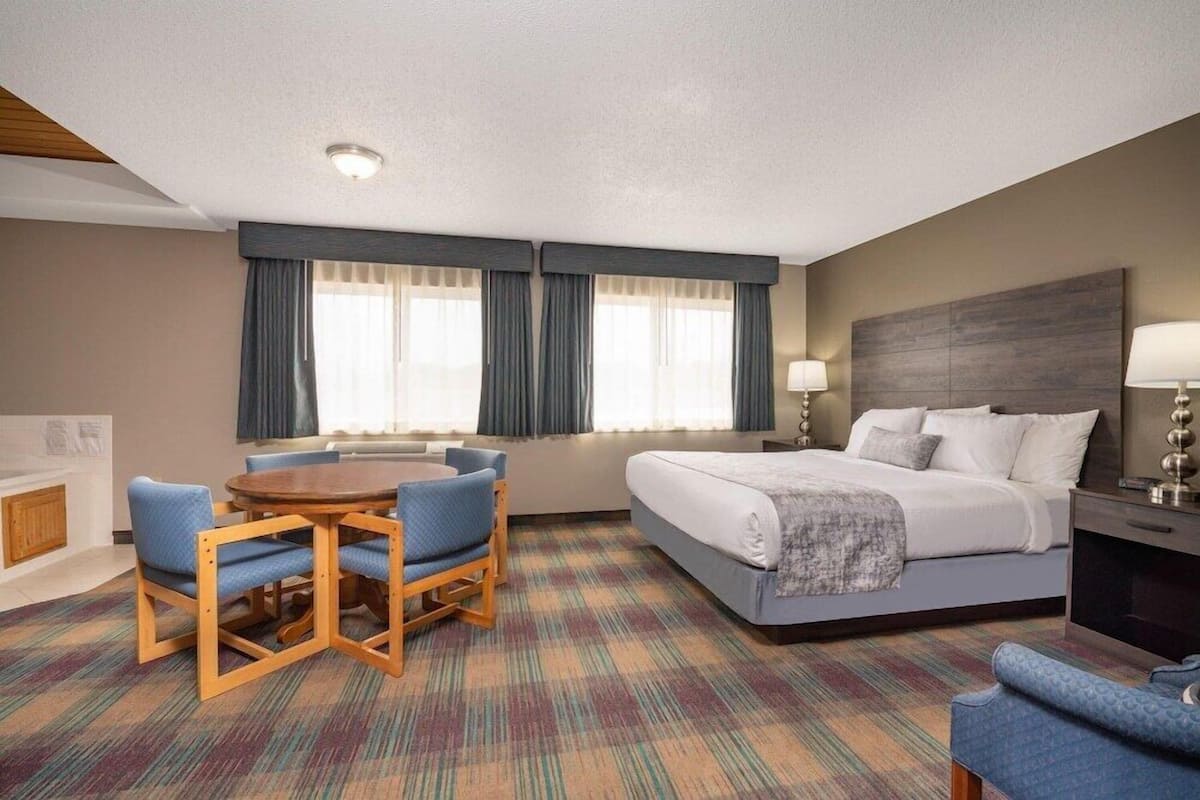 A hotel room with a bed, a round table with chairs, a desk with a lamp, and a view of the outside through large windows.