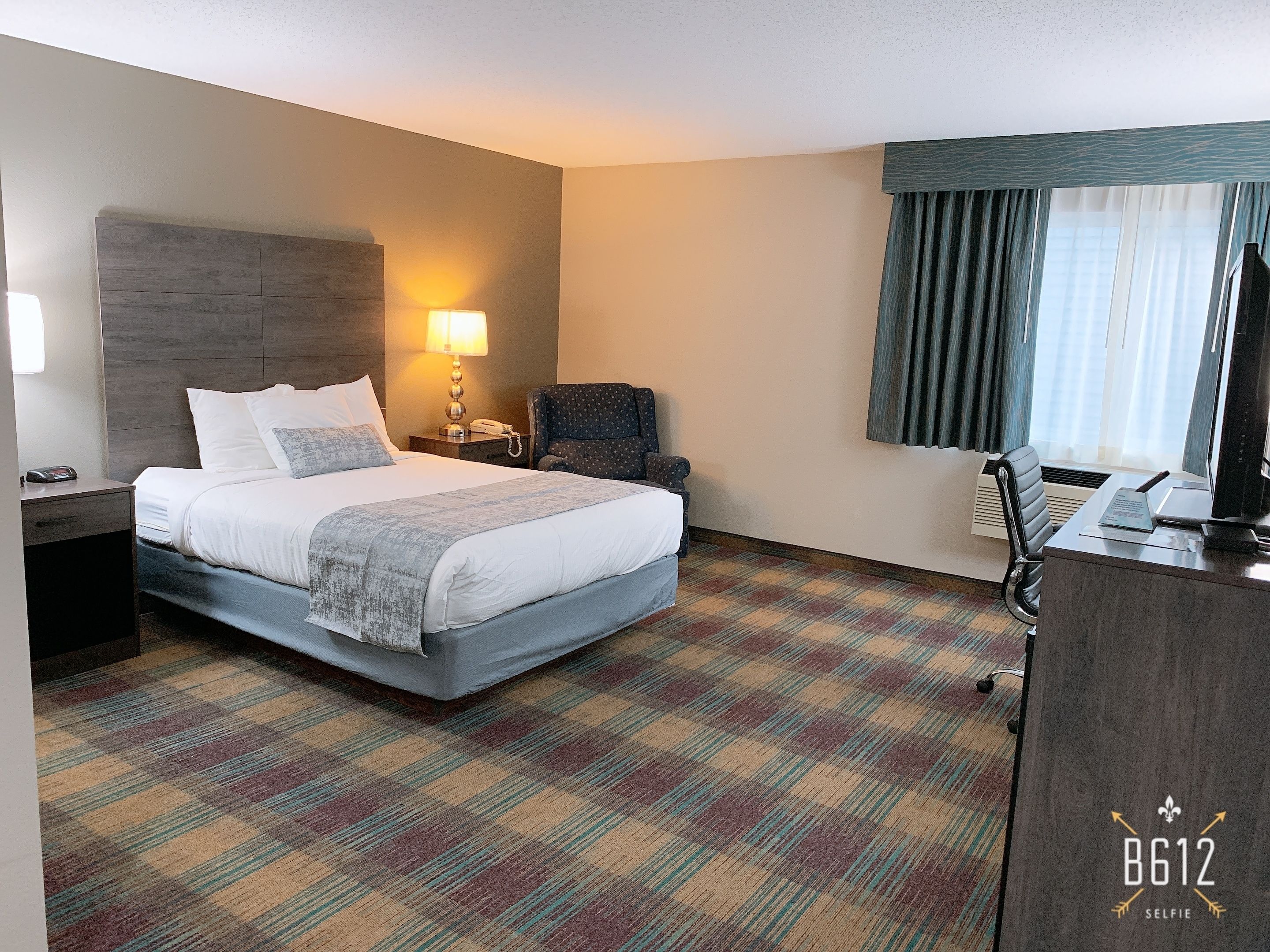 Una habitación de hotel con cama, escritorio, silla, lámpara y ventana con cortinas.