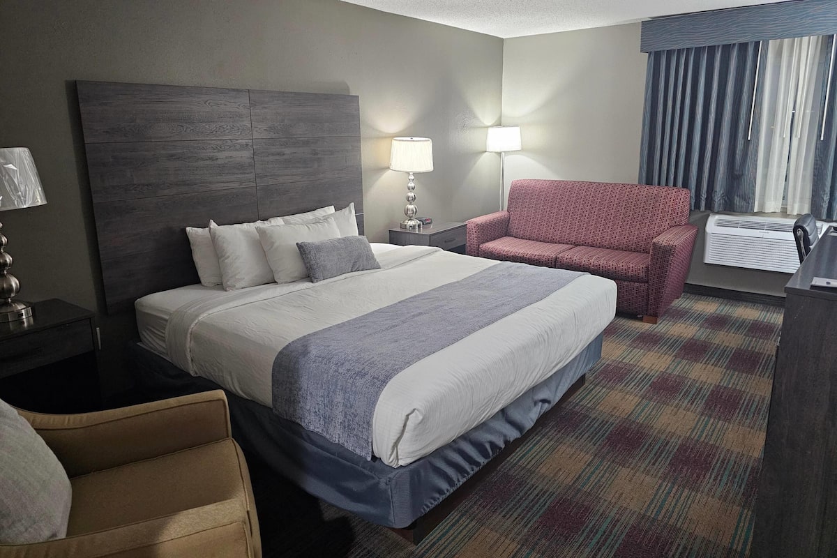 A hotel room with a large bed, two bedside lamps, a red armchair, a desk with a lamp, and a window with curtains.