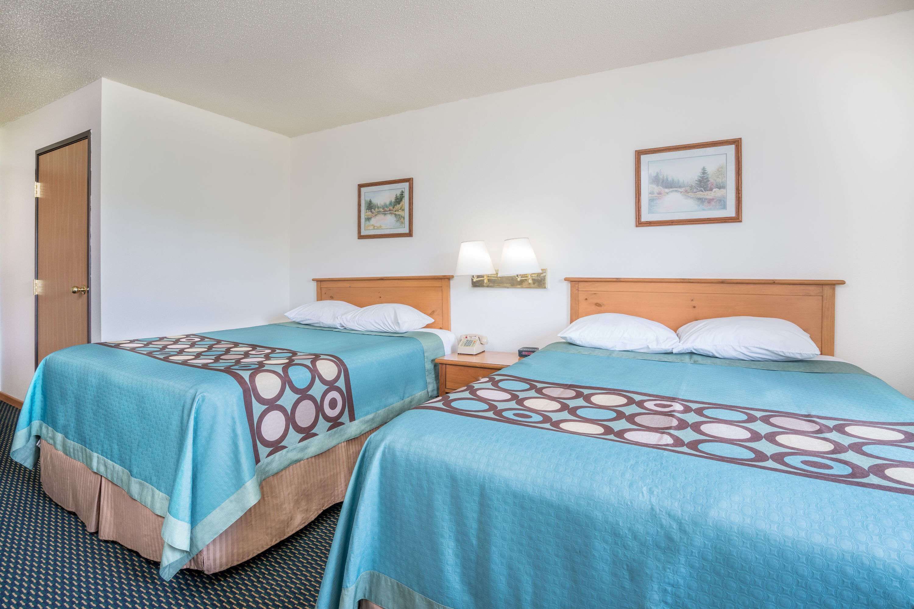 A hotel room with two beds, a door, a nightstand with a lamp, and two framed pictures on the wall.