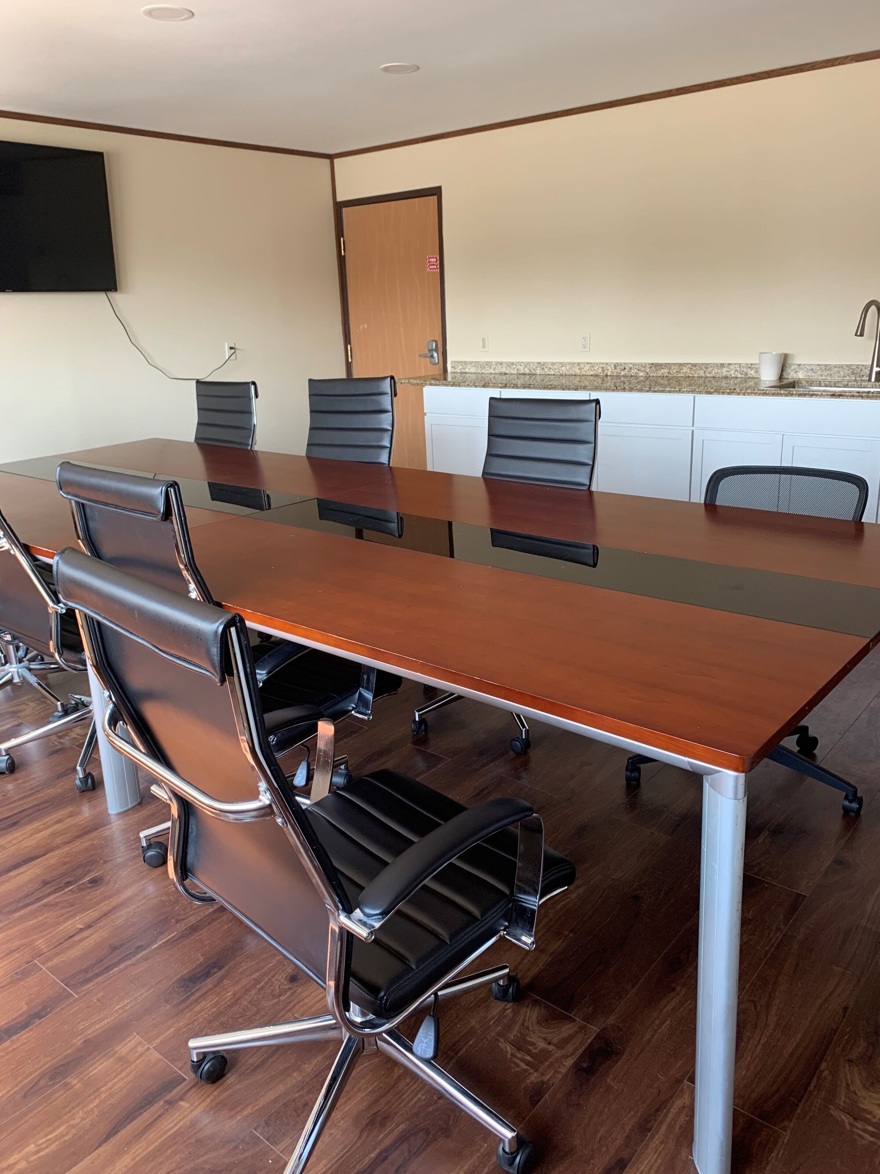 A conference room with a long wooden table, black leather chairs, and a white kitchenette in the background.