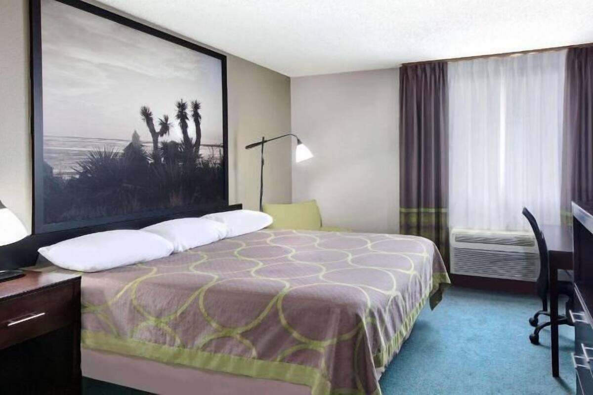A hotel room with a bed, a desk, a chair, a desk lamp, a large window with curtains, and a wall-mounted TV.