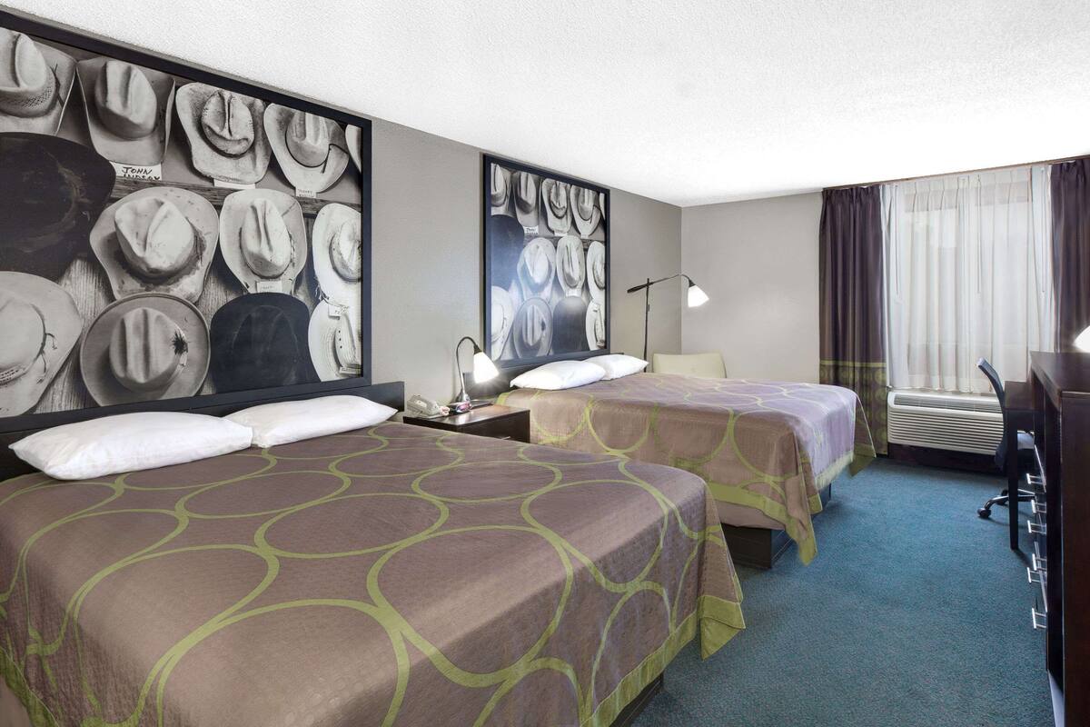 A hotel room with two beds, a desk, a chair, a lamp, and a large wall art of cowboy hats.