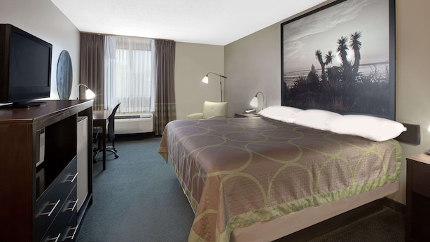 Une chambre d’hôtel comprenant un lit, un bureau avec une chaise, une télévision, une grande fenêtre avec des rideaux et un tableau au mur.