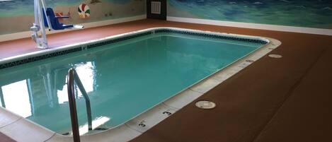 Una piscina interior con un mural de una escena de playa, avisos del socorrista y normas de la piscina expuestas en la pared.