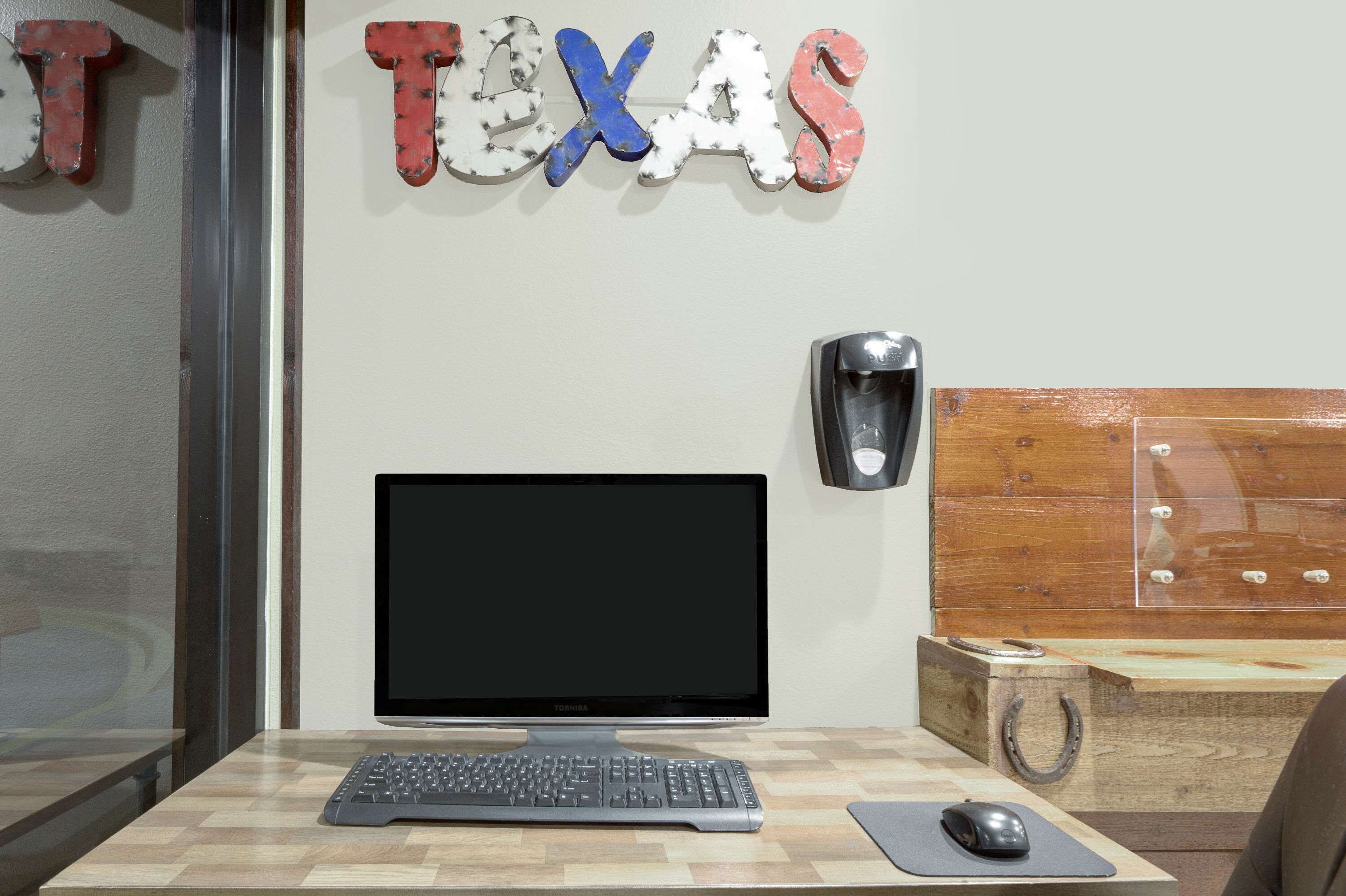 A workspace with a computer monitor, keyboard, and mouse on a wooden desk. A wall-mounted speaker is visible, and a decorative sign spelling "TEXAS" is above the monitor.