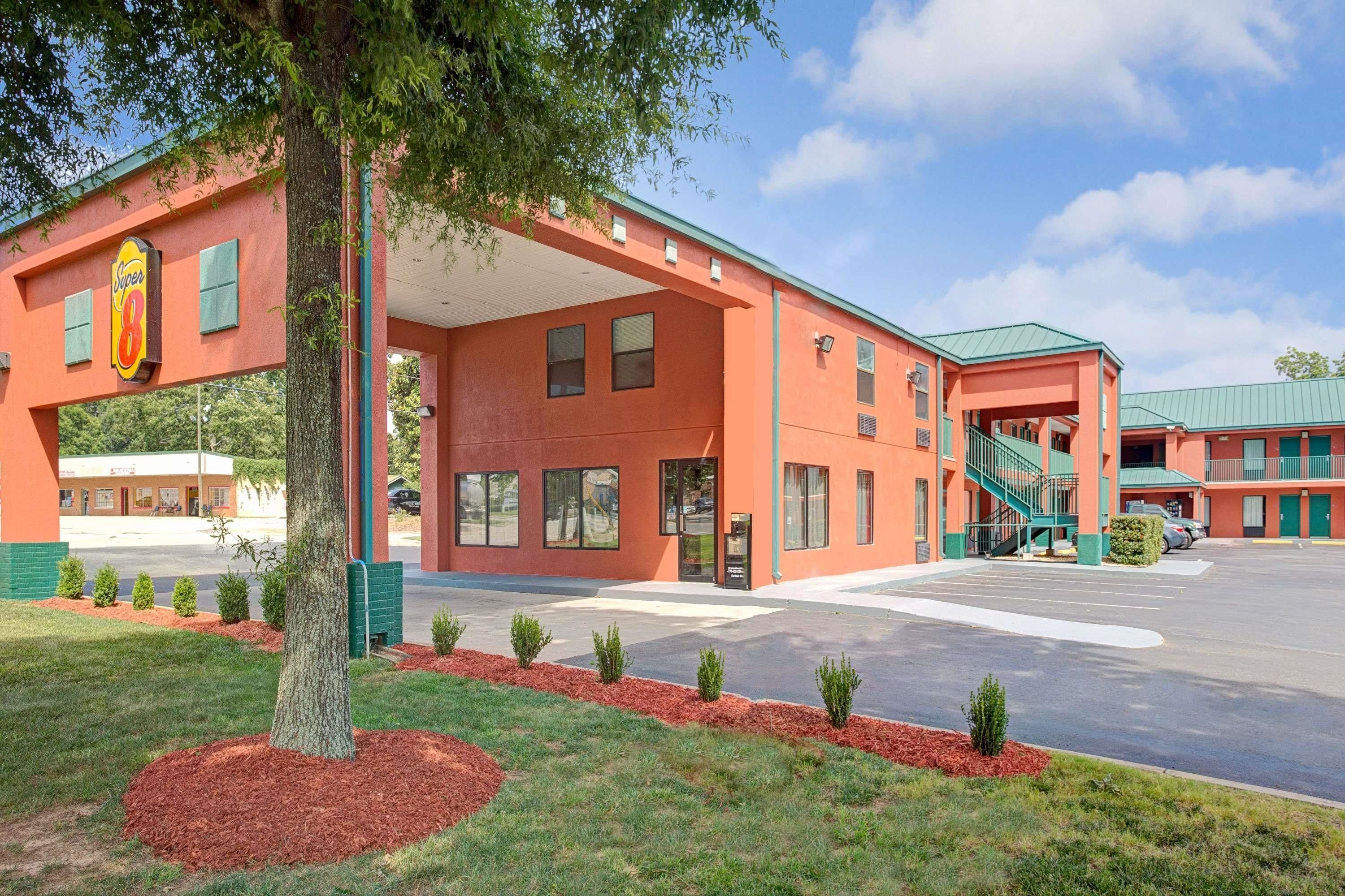 A motel with a red exterior, a covered parking area, and a small garden with shrubs.