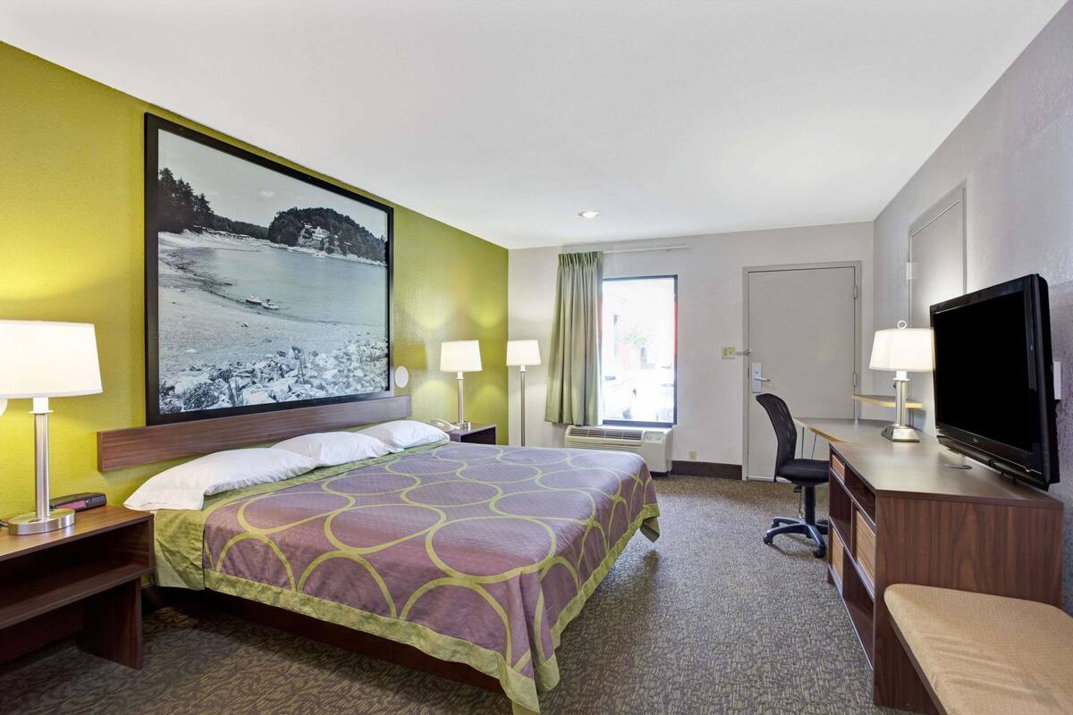 A hotel room with a bed, two bedside lamps, a flat-screen TV, a desk with a chair, and a large window with curtains.