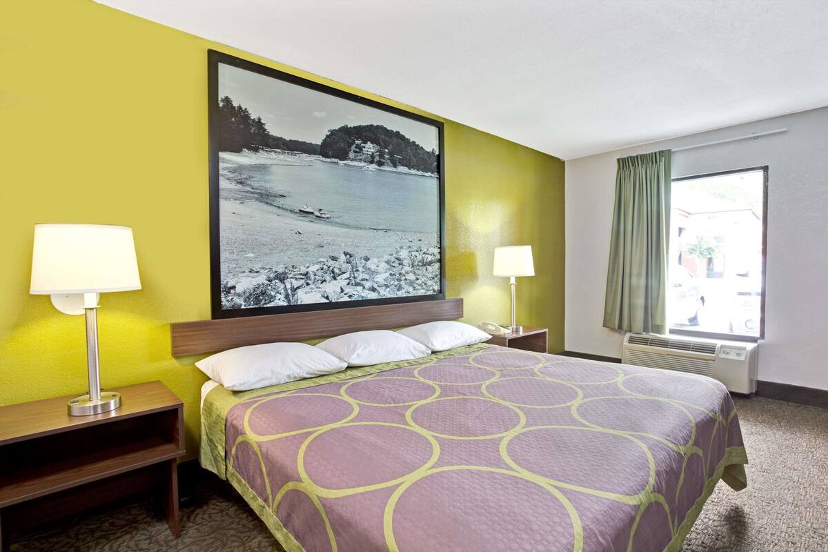 A hotel room with a bed, two bedside lamps, a large framed picture on the wall, a window with a view, and a heater.