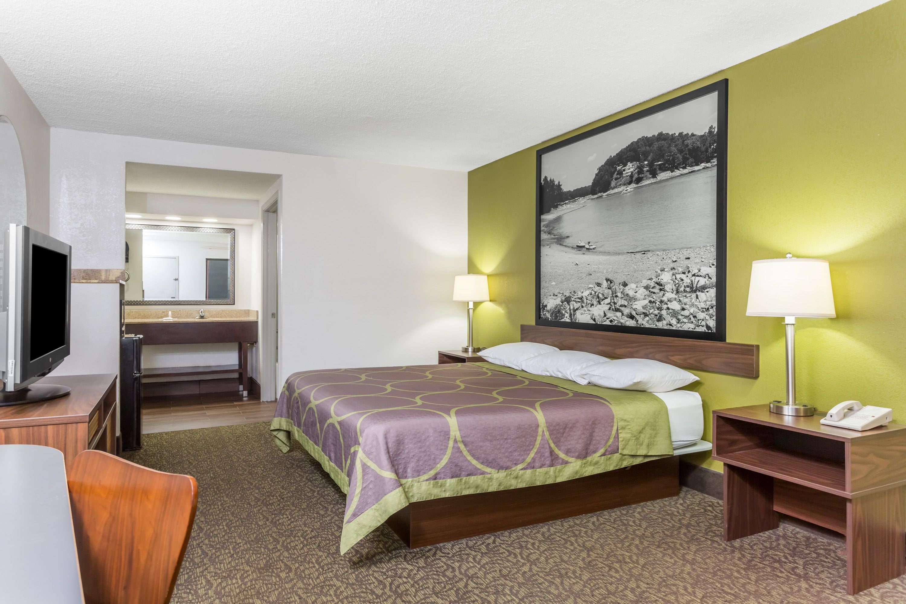 A hotel room with a bed, bedside lamps, a desk with a television, a bathroom with a sink and mirror, and a large framed picture on the wall.