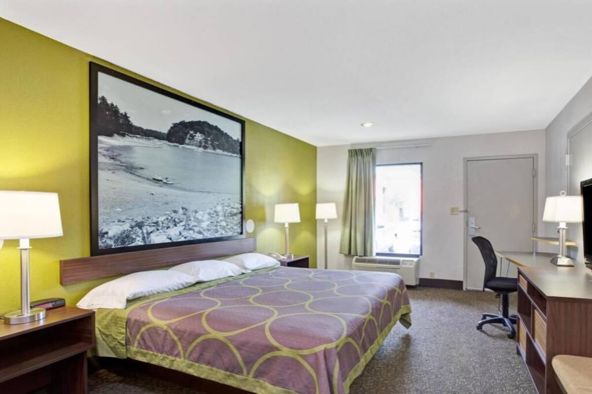 A hotel room with a bed, two bedside lamps, a desk with a chair, a large window, and a painting on the wall.