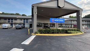 A motel with a 'Now Open' sign, a covered parking area, and a building with a grey roof and multiple windows. - Motel 6 Charlotte, NC - Northlake (Charlotte)