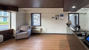 A reception area with a wooden floor, a grey sofa, a window with a view of parked cars, and a reception desk with a computer and a small plant. - Motel 6 Charlotte, NC - Northlake (Charlotte)