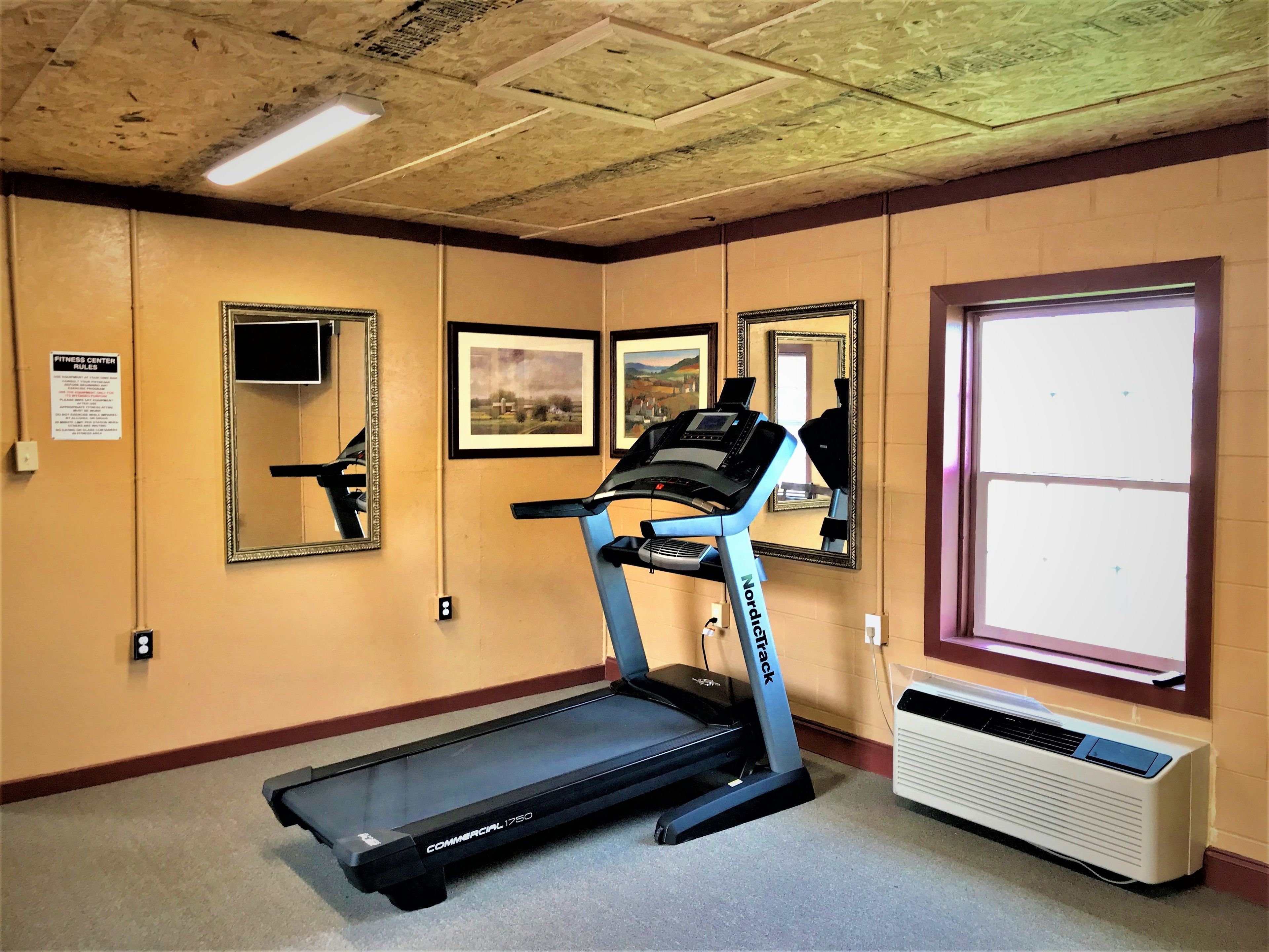 A fitness center with a treadmill, mirrors, framed pictures, and an air conditioning unit.